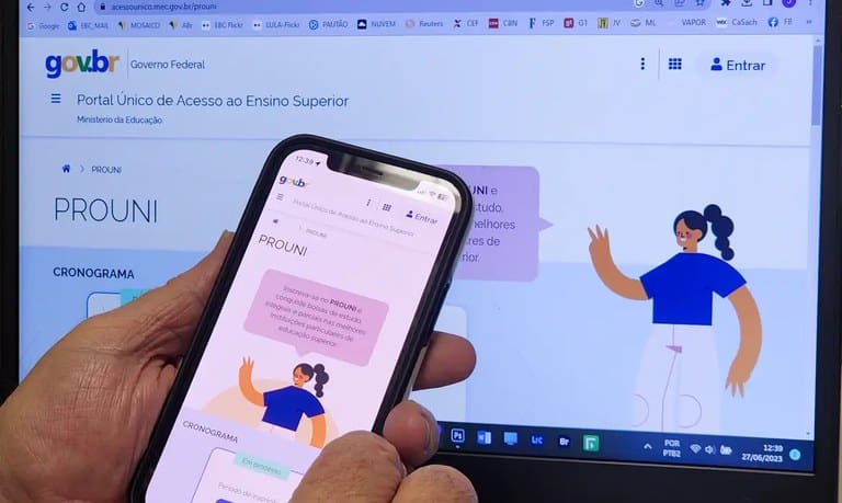 Em plano de fundo, o site do Prouni está aberto em uma tela de computador. Em primeiro plano, duas mãos seguram um celular, onde também está aberto o site do Prouni.