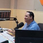 Um homem branco tem cabelo preto e usa camisa azul. Ele fala em um microfone preto, que está à sua frente. Há uma mesa de som e três monitores, dos quais é possível ver a tela de dois. A parede ao fundo é amarela.