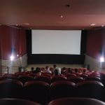 Uma sala de cinema é vista da última fileira. Ainda não há exibição, e algumas pessoas já estão nos assentos.