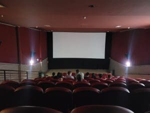 Uma sala de cinema é vista da última fileira. Ainda não há exibição, e algumas pessoas já estão nos assentos.