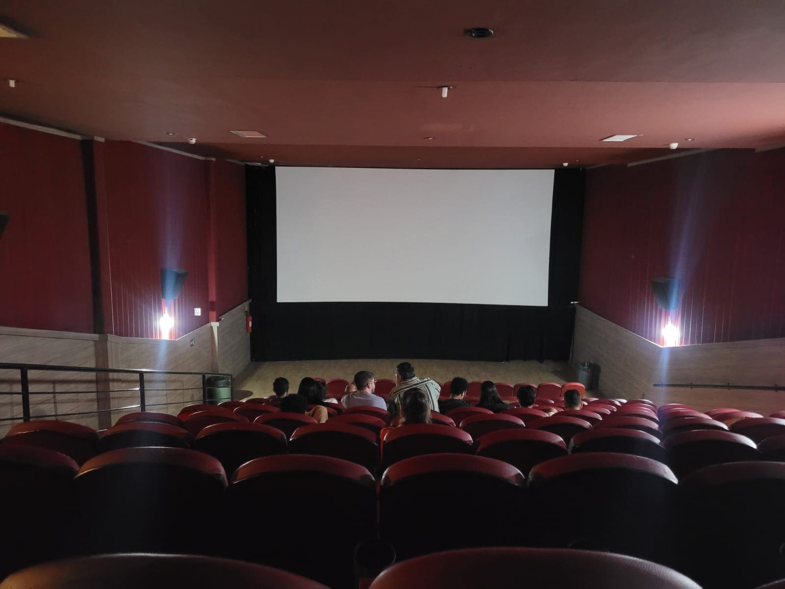 Uma sala de cinema é vista da última fileira. Ainda não há exibição, e algumas pessoas já estão nos assentos.