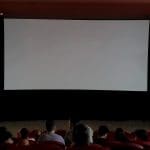 A foto é tirada das últimas fileiras de uma sala de cinema, que está com poucas luzes ligadas. A grande tela branca ainda não exibe nenhum filme. Há algumas pessoas na sala.