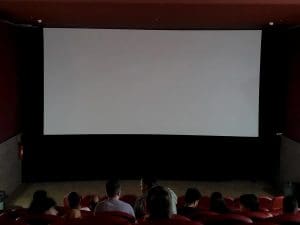 A foto é tirada das últimas fileiras de uma sala de cinema, que está com poucas luzes ligadas. A grande tela branca ainda não exibe nenhum filme. Há algumas pessoas na sala.