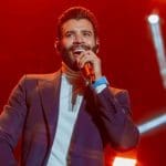 O cantor Gusttavo Lima, um homem branco, adulto, de cabelo e barba pretos, canta segurando um microfone. Ele usa camisa branca e blazer preto. Ao fundo, as luzes estão vermelhas.