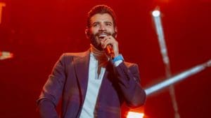 O cantor Gusttavo Lima, um homem branco, adulto, de cabelo e barba pretos, canta segurando um microfone. Ele usa camisa branca e blazer preto. Ao fundo, as luzes estão vermelhas.