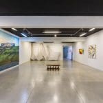Uma sala de galeria tem as paredes brancas. É possível ver três delas. À esquerda, uma pintura que ocupa toda a altura da parede. Ao fundo, esculturas de isopor e ferro. À direita, alguns quadros. No meio, um banco que também compõe a exposição.