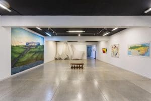 Uma sala de galeria tem as paredes brancas. É possível ver três delas. À esquerda, uma pintura que ocupa toda a altura da parede. Ao fundo, esculturas de isopor e ferro. À direita, alguns quadros. No meio, um banco que também compõe a exposição.