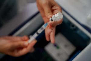 Duas mãos seguram uma seringa e um recipiente onde está uma dose de vacina. A seringa puxa o conteúdo do recipiente.