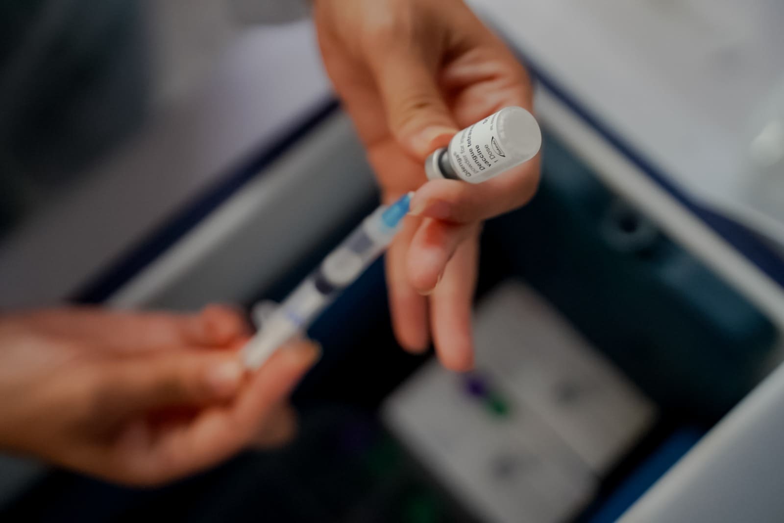 Duas mãos seguram uma seringa e um recipiente onde está uma dose de vacina. A seringa puxa o conteúdo do recipiente.