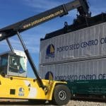Na imagem, um trator amarelo levanta dois contêineres brancos. Neles, está escrito "Porto Seco Centro Oeste". É dia, e o céu está azul.