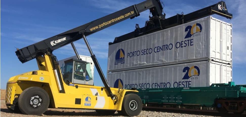 Na imagem, um trator amarelo levanta dois contêineres brancos. Neles, está escrito "Porto Seco Centro Oeste". É dia, e o céu está azul.