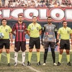 Árbitros, o capitão do Anápolis e o capitão do Iguatu estão alinhados antes da partida começar. Ao fundo, uma faixa com a palavra "tricolor" e a plateia.