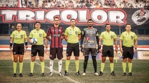 Árbitros, o capitão do Anápolis e o capitão do Iguatu estão alinhados antes da partida começar. Ao fundo, uma faixa com a palavra "tricolor" e a plateia.