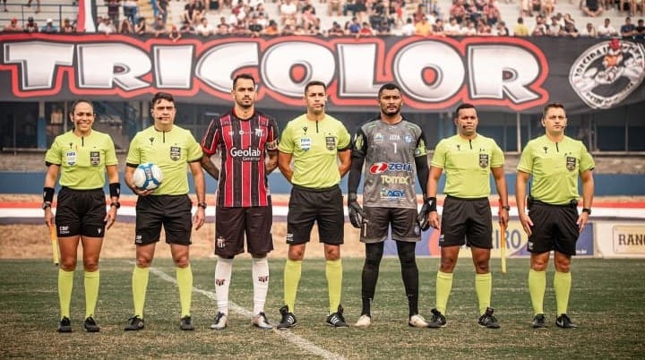 Árbitros, o capitão do Anápolis e o capitão do Iguatu estão alinhados antes da partida começar. Ao fundo, uma faixa com a palavra "tricolor" e a plateia.