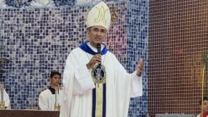 Um bispo veste as roupas características de seu posto religioso e segura um microfone, enquanto ministra um culto.