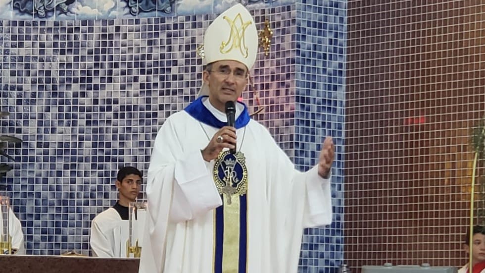 Um bispo veste as roupas características de seu posto religioso e segura um microfone, enquanto ministra um culto.