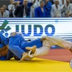 Dois judocas se enfrentam no tatame. Uma placa ao fundo diz "Judô", atrás da qual aparece a plateia.