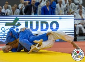 Dois judocas se enfrentam no tatame. Uma placa ao fundo diz "Judô", atrás da qual aparece a plateia.