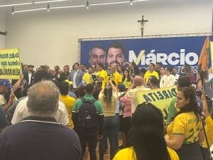 Várias pessoas estão em um palanque durante uma sessão na Câmara Municipal. Ao fundo, um banner com o rosto de Márcio Corrêa,
