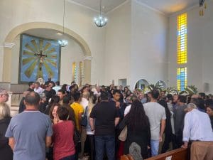 Diversas pessoas estão reunidas em uma igreja para o velório de Dom Dilmo. É possível ver coroas de flores ao fundo.
