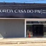 Fachada da Igreja Casa do Pai, com as paredes cinzas. A placa é preta, e o nome está escrito em letras brancas. As portas estão abertas.