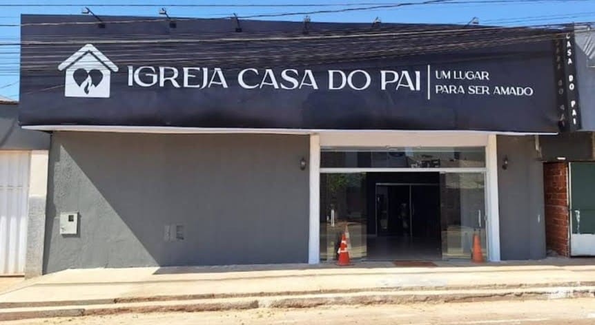 Fachada da Igreja Casa do Pai, com as paredes cinzas. A placa é preta, e o nome está escrito em letras brancas. As portas estão abertas.
