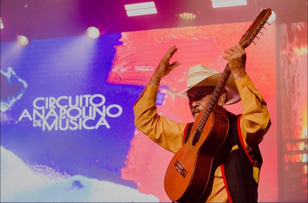 Um violeiro toca em frente ao telão que diz "Circuito Anapolino de Música".