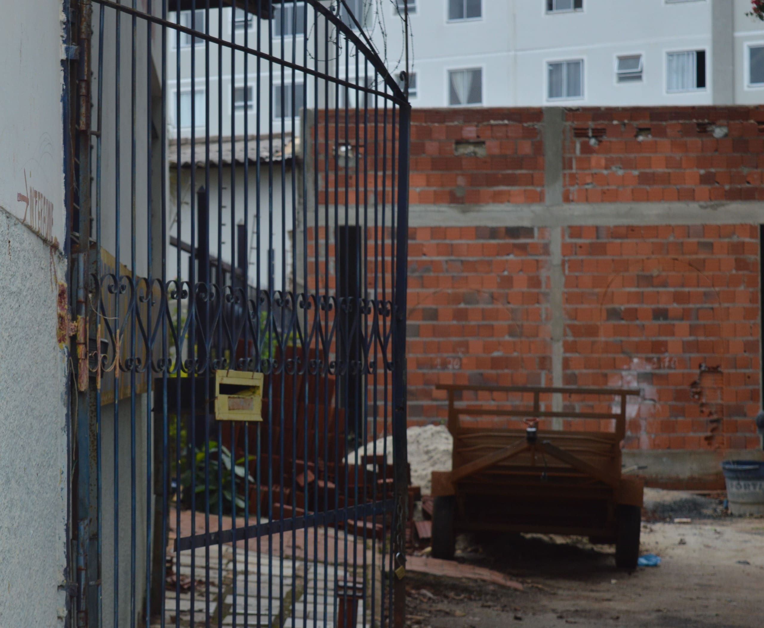Local é visto do lado de fora. Há um portão aberto em primeiro plano. Ao fundo, uma carretinha, um monte de areia e uma casa de tijolos que ainda não foi rebocada.