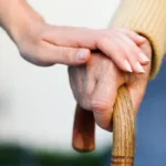Imagem mão de idoso segurando bengala e uma mão jovem por cima