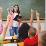 Imagem professora em sala de aula, alunos aparecem erguendo a mão, a frente está professora segurando um livro