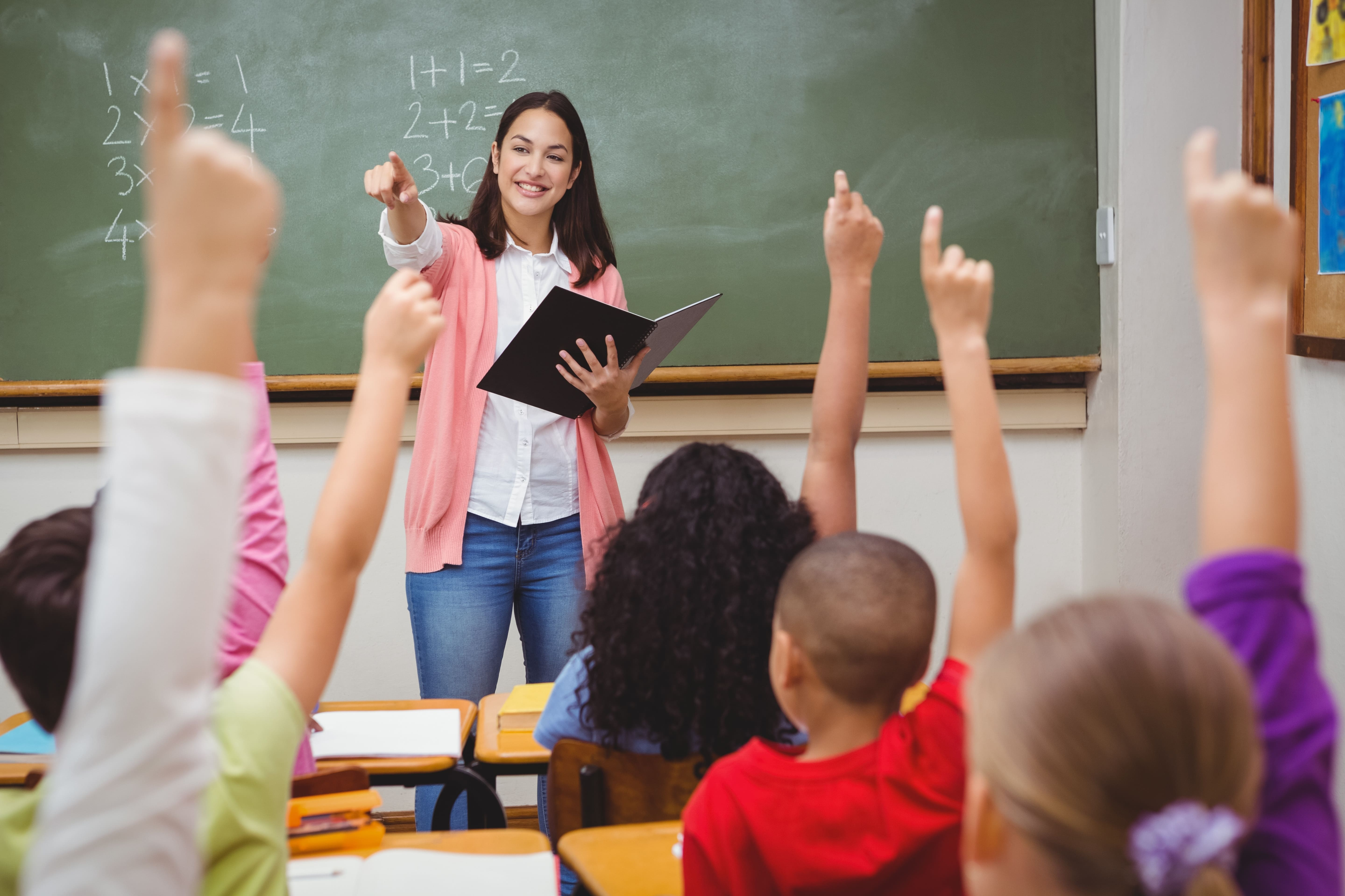 Imagem professora em sala de aula, alunos aparecem erguendo a mão, a frente está professora segurando um livro