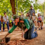 Imagem Mulher ajoelhada plantando árvore, crianças em volta assistem o plantio