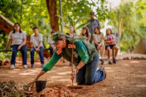 Imagem Mulher ajoelhada plantando árvore, crianças em volta assistem o plantio