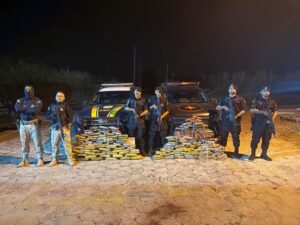 Imagem policiais militares e da PRF segurando armas ao lado de 200kg de drogas apreendidas