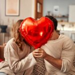 Imagem casal sentados com um balão em formato de coração vermelho cobrindo o rosto de ambos