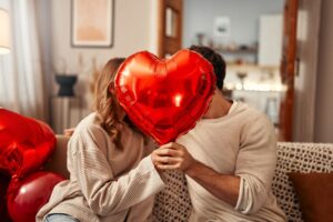 Imagem casal sentados com um balão em formato de coração vermelho cobrindo o rosto de ambos