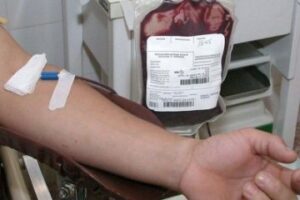 Imagem Doação de Sangue. Cateter está retirando sangue de uma pessoas, bolsa de sangue aparece cheia ao fundo.