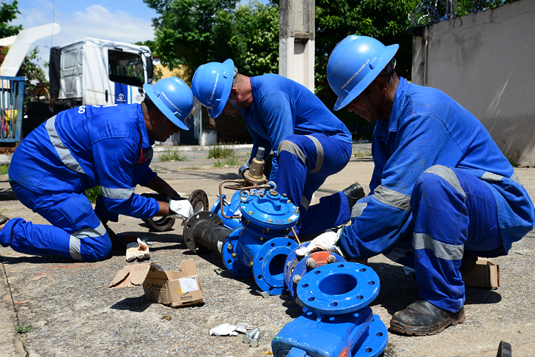 Imagem equipes da Saneago realizam manutenção. Hoemns com capacete estão trabalhando com canos e uma rua