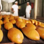 Imagem pão no forno para assar, ao fundo aparece duas pessoas de branco