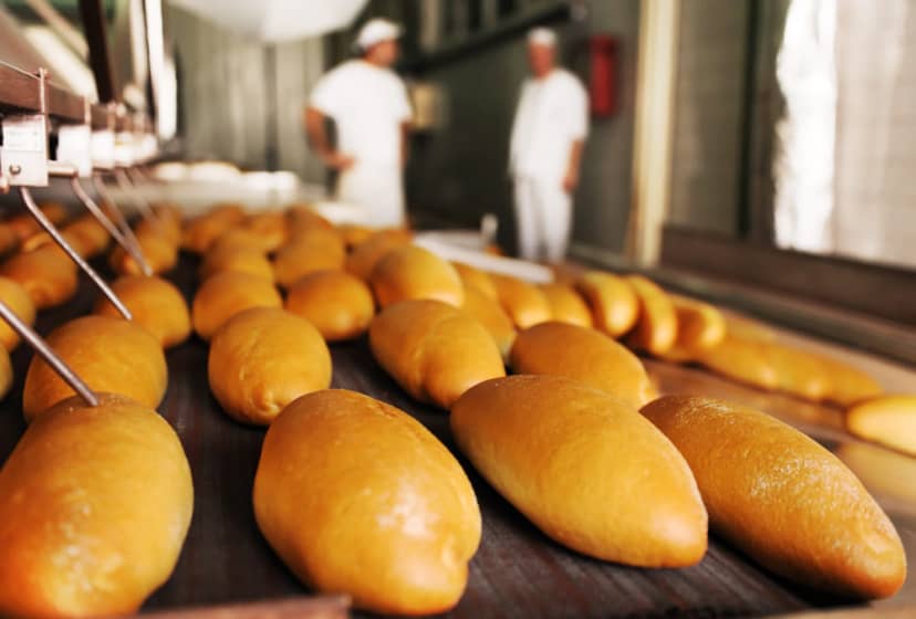 Imagem pão no forno para assar, ao fundo aparece duas pessoas de branco