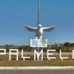 Imagem letreiro na entrada da cidade com o nome PALMELO em cima uma placa e uma pomba branca