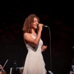 Imagem mulher cantando, com microfone próximo a boca, ela veste vestido branco