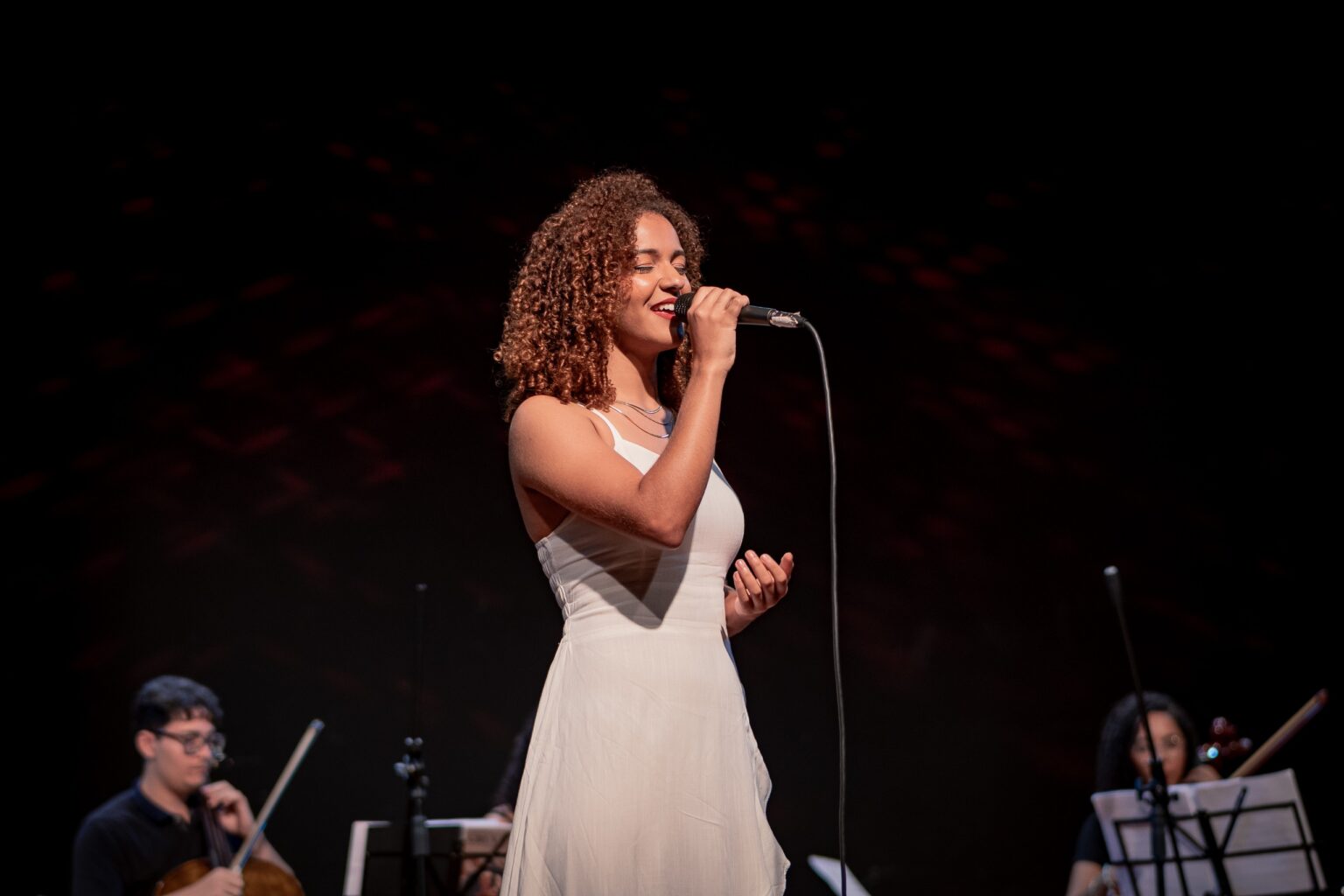Imagem mulher cantando, com microfone próximo a boca, ela veste vestido branco
