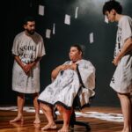 Imagem espetáculo “Página Um” um homem ao centro sentado na cadeiras e dois em pé