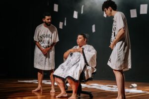 Imagem espetáculo “Página Um” um homem ao centro sentado na cadeiras e dois em pé