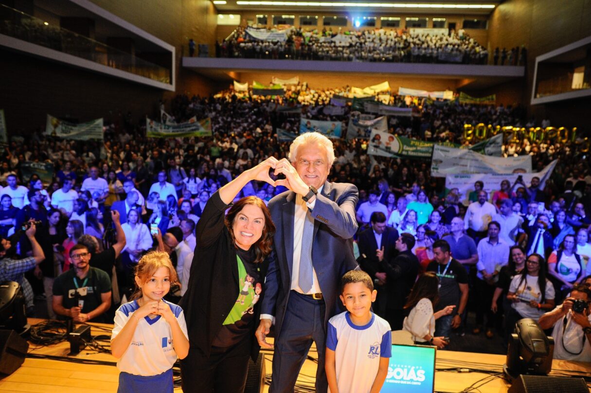 Imagem governador Ronaldo Caiado e secretaria de Educação Fátima Gavioli. ao lado de duas crianças, ao fundo alunos de rede estadual de educação.