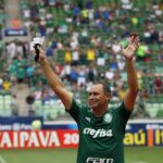 Imagem Evair Paulino de mãos erguidas, ele está em um estádio, vestindo a camisa do Palmeiras e segurando um microfone
