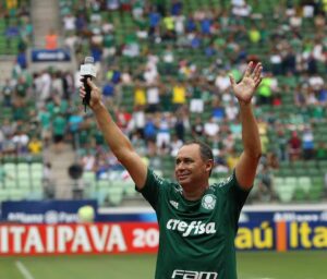 Imagem Evair Paulino de mãos erguidas, ele está em um estádio, vestindo a camisa do Palmeiras e segurando um microfone