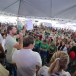 Imagem vários pessoas em evento da Alego, ao centro um homem fala com microfone na mão