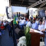 Imagem Daniel Vilela falando ao microfone, pessoas aparecem ao fundo em cima de palco